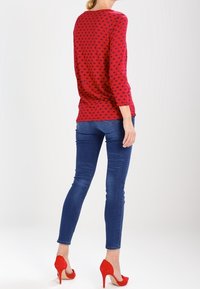 Maglietta a maniche lunghe a fantasia rossa con jeans skinny blu e tacchi alti rossi. La maglietta presenta piccoli pois blu scuro su uno sfondo rosso.