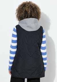 Chaleco negro acolchado con capucha gris, con costuras en forma de chevron, superpuesto sobre una camisa de manga larga a rayas azul y blanca.