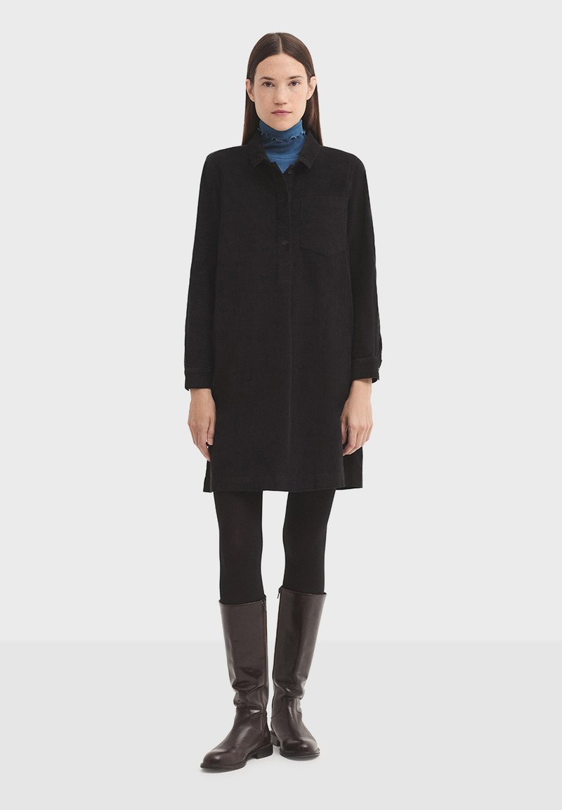 Čierne semišové šaty košeľového strihu s prednou vreckom, gombíkovým golierom, dlhými rukávmi a voľným strihom, skombinované s čiernymi legínami a hnedými vysokými topánkami po kolená.