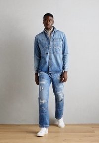 Chaqueta de mezclilla azul claro con dos bolsillos en el pecho y una camisa debajo, combinada con jeans desgastados de mezclilla azul claro y zapatillas blancas.