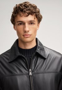 Giubbotto di pelle nera con una texture liscia, dotato di chiusura a cerniera e colletto classico. Indossato sopra un maglione di lana blu navy.