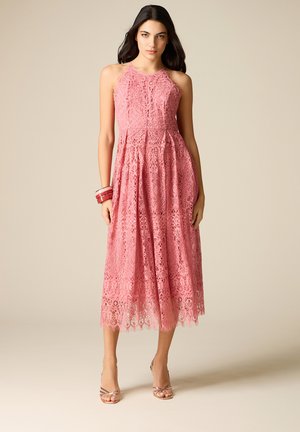 Vrouw draagt een mouwloze roze kanten midi-jurk met geplooide rok, staand met ontspannen handen langs het lichaam, beige achtergrond.