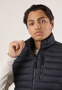 Giovane uomo con capelli scuri che sistema il collo di un gilet nero trapuntato con cerniera sopra una camicia a maniche lunghe beige chiaro, sullo sfondo neutro.