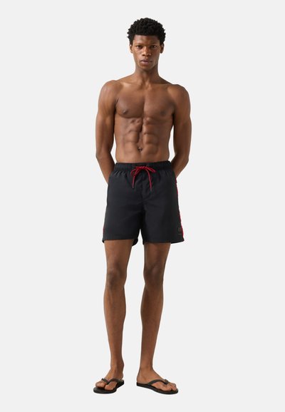 Homme torse nu debout, les mains derrière le dos, portant un short de bain noir avec un cordon rouge et des tongs noires, sur un fond uni.