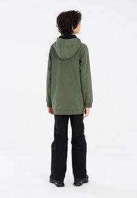Groene hoodie-jack met elastische manchetten, gecombineerd met zwarte broek en stevige zwarte laarzen, gezien van achteraf tegen een effen achtergrond.