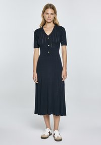 Vestito a maglia blu navy caratterizzato da un collo a V, maniche corte, chiusura con bottoni, vita a coste e gonna svasata con dettagli in punto occhiello; indossato con sneakers.