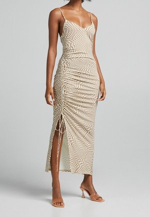 Beige midi-kjole med slim pasform og brun prikket mønster, rynket sideslids bundet med snore, båret med klare hælede sandaletter med åben tå.