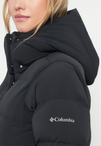 Närbild på en kvinna som bär en svart Columbia-dunjacka med en stor huva och synlig logotyp på vänster ärm.