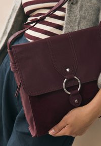 Handtas van bordeauxrood leer en suède met een klep, zilverkleurige metalen ring en verstelbare riem, vastgehouden in de hand van een persoon.