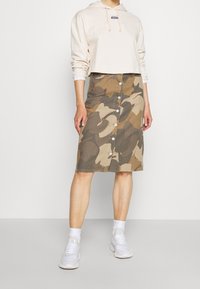 Kamouflage-mönstrad denimkjol med framknappar och en figurnära silhuett, ihop med en kort beige huvtröja och vita sneakers.