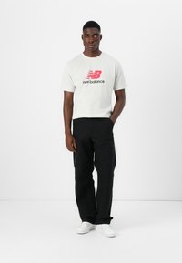 Gray short-sleeve t-shirt with pink New Balance logo, paired with black pants and white sneakers. Plain background.