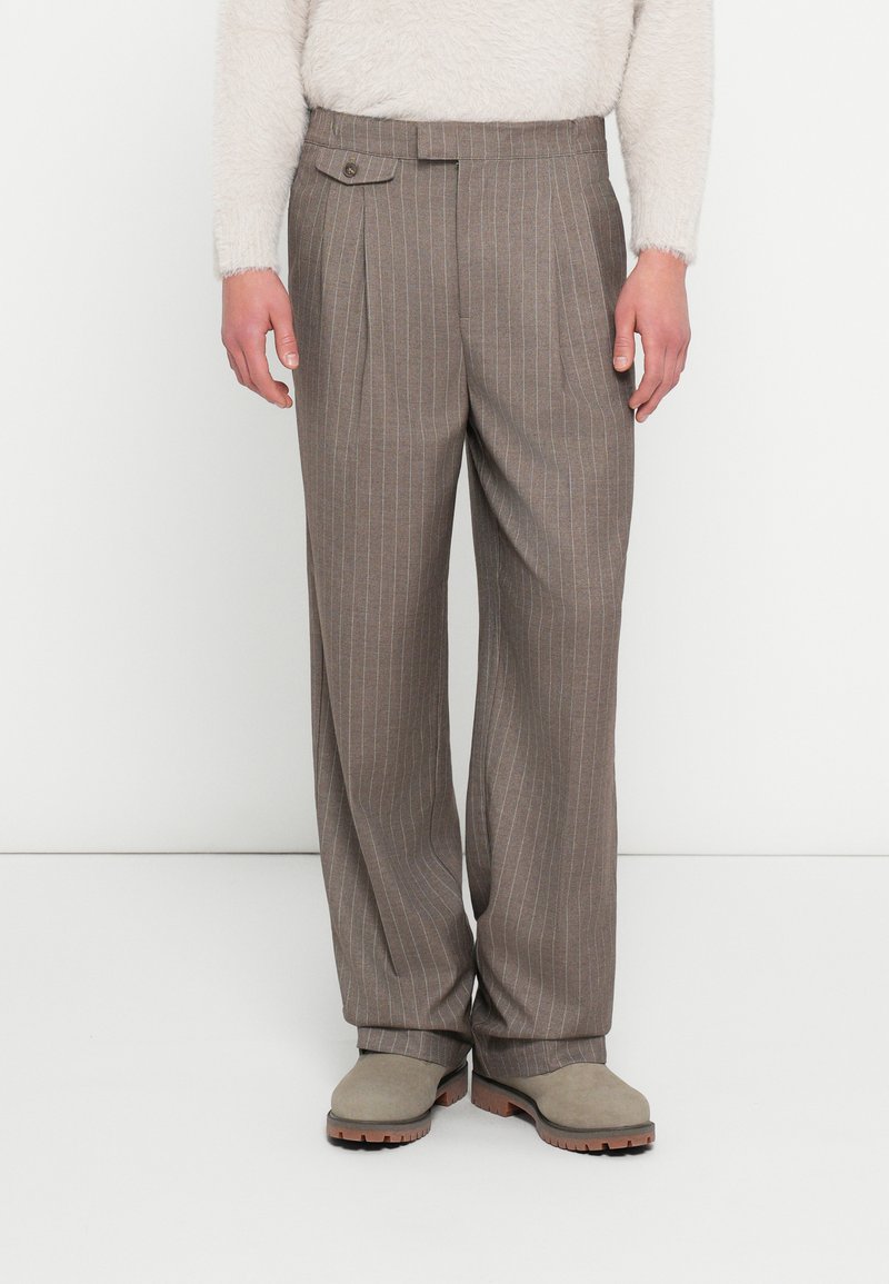 Hnedé nohavice z vlnenej zmesi s modrými prúžkami, s vreckom so zapínaním na gombík a záhybmi. Nosia sa so sivými slip-on topánkami.