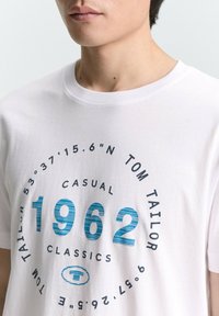 Valkoinen puuvillainen T-paita, jossa sininen pyöreä graafinen kuvio, jossa lukee "1962" raidoitetussa muotoilussa, ympärillä teksti ja logo. Klassinen pyöreä kaula-aukko.