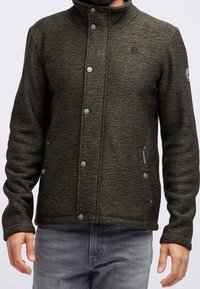 Uomo che indossa una giacca in maglia verde scuro con zip e bottoni a pressione, due tasche frontali e un logo sul petto e sulla manica.