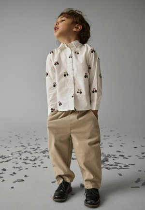 Jeune enfant portant un t-shirt blanc à motif tracteur, un pantalon beige et des chaussures noires, debout sur un sol gris éclaboussé de morceaux de papier.
