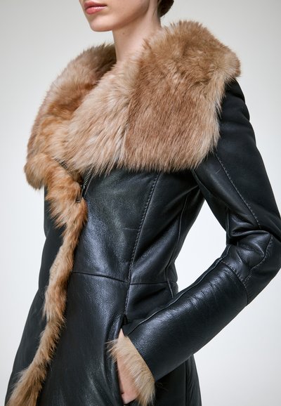 Personne portant un manteau en cuir noir ajusté avec un large col en fourrure marron et des bordures en fourrure sur les manches, main dans la poche du manteau.