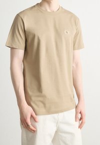 Kamelfärgad bomulls-T-shirt med rundad hals, korta ärmar och en liten broderad logotyp på vänster bröst. Slät textur, avslappnad passform.