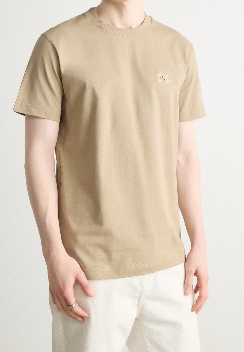 Kamelfärgad bomulls-T-shirt med rundad hals, korta ärmar och en liten broderad logotyp på vänster bröst. Slät textur, avslappnad passform.
