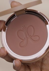 Compact redondo con un acabado en oro rosa, que presenta un diseño de mariposa sobre una superficie de polvo prensado suave y marrón cálido.