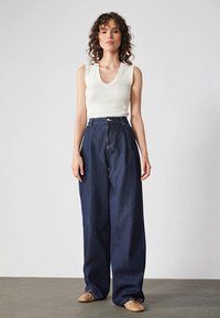 Kvinna med lockigt hår som bär en vit ärmlös topp, högmidjade vida mörkjeans och beige balettklackar, som står mot en enfärgad vägg.