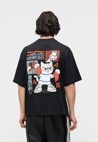 Άτομο που φοράει μαύρο μπλουζάκι Adidas με καρτούν γάτα που σπάει μια καρδιά και ιαπωνικό κείμενο στην πλάτη.