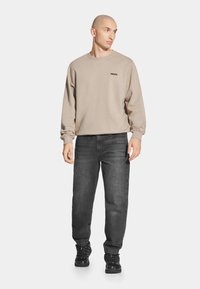 Beżowa bluza z okrągłym dekoltem i czarnym logo, zestawiona z ciemnoszarymi spodniami i czarnymi trampkami. Model stoi w neutralnej pozycji.