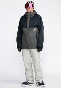 Sneeuwjas in donkergrijs en zwart, met een ritstas, gecombineerd met lichtgrijze sneeuwbroek en zwart-witte sneeuwschoenen. Voorzien van een skibril en handschoenen.