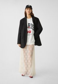 Černý blazer přes bílou grafickou mikinu s potiskem třešní, kombinovaný s dlouhou krajkovou sukní v krémové barvě a červenými podpatky. Na hlavě černá kšiltovka.