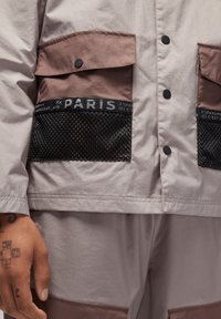 Lichtgrijze jas met bruine accenten, met twee zakken, zwart gaasdetail en drukknoopsluitingen. De stof heeft een gladde textuur.