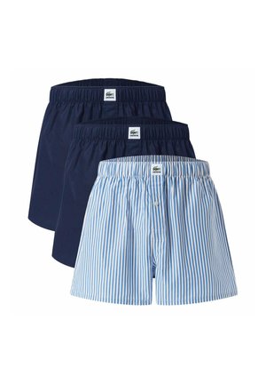 Tres pares de calzoncillos tipo bóxer para hombre de Lacoste apilados, dos lisos en azul marino y uno a rayas azul claro y blanco con bragueta con botones.