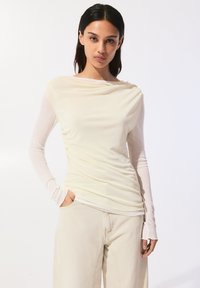 Långärmad topp med rynkad design i ljus krämfärg, lager på lager över en åtsittande bas. Det mjuka tyget faller elegant och kombineras med neutrala beige byxor.