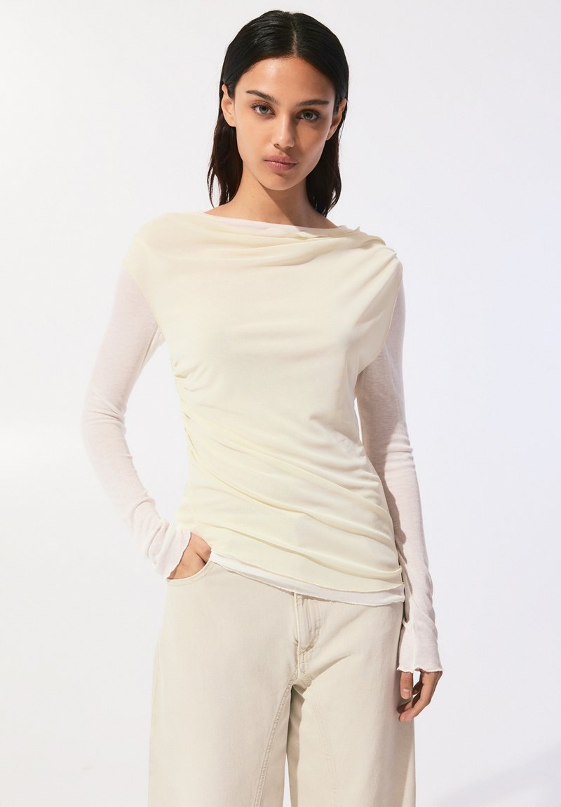 Långärmad topp med rynkad design i ljus krämfärg, lager på lager över en åtsittande bas. Det mjuka tyget faller elegant och kombineras med neutrala beige byxor.