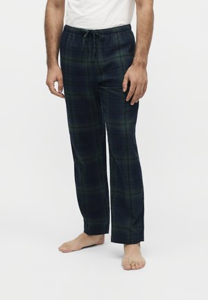 Homme portant un pantalon de pyjama à carreaux bleu foncé et vert avec cordon de serrage et une chemise blanche, debout pieds nus devant un fond uni.
