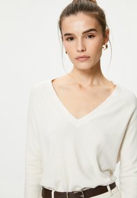 Biały sweter w serek wykonany z miękkiego materiału, z długimi rękawami i luźnym krojem, zestawiony z brązowym paskiem w talii.