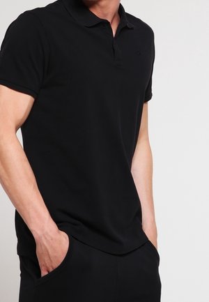 Camiseta tipo polo negra de algodón. Mangas cortas, cuello y ajuste holgado. Logo sutil en el pecho. Se lleva con las manos en los bolsillos.