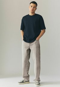 Marineblauwe katoenen T-shirt met korte mouwen, gecombineerd met lichtgrijze denim jeans en zwart-witte sneakers. Model staat met de handen in de zakken.