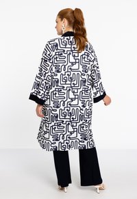 Lang, witte kimono met een zwart abstract patroon, driekwartmouwen en zwarte afwerking, gecombineerd met donkere broeken.