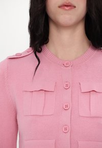 Femme portant un cardigan rose à boutons avec des poches à rabat et des épaulettes boutonnées, vue du cou à la poitrine.