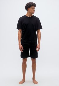 Νέος άνδρας με μαύρο μπλουζάκι και σορτς, όρθιος ξυπόλητος σε λευκό φόντο, κοιτάζει πλάγια με χαλαρή στάση.