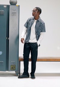 Ριγέ πουκάμισο με κοντό μανίκι πάνω από λευκό πουκάμισο με μακρύ μανίκι, μαύρο παντελόνι και μαύρα αθλητικά παπούτσια. Γκρι ντουλάπα και ξύλινο παγκάκι στο φόντο.