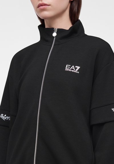 Жена, носеща черно яке с цип с висока яка и бяло лого EA7 Emporio Armani на лявата част на гърдите.