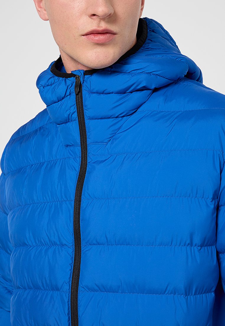 Giacca a vento blu con zip frontale, cappuccio regolabile e motivo a righe cucite orizzontali. Accent del zip nera che aggiunge contrasto.