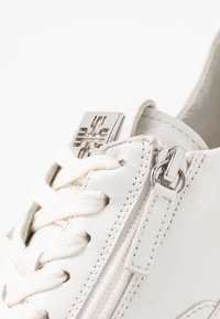 Valkoinen nahkainen lenkkari, jossa on nyöritys edessä, hopeiset metalliosat, sivuvetoketju ja nyörien läheisyydessä oleva teksturoitu pinta.