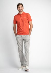 Oranje polo shirt met een kraag, grijze broek en witte sneakers. Het shirt heeft een logo aan de linkerkant en een zachte textuur.