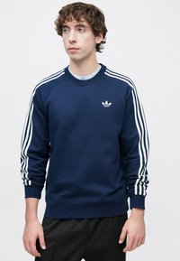 Μπλε ναυτικό φούτερ με τρεις λευκές ρίγες σε κάθε ώμο, στρογγυλή λαιμόκοψη και μικρό λευκό λογότυπο στο στήθος. Υλικό από βαμβακερό μείγμα.