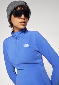 Plavi flis pulover s ovratnikom na zip, s bijelim logom na prsima. Dodaci uključuju sivi pleteni beanie i crne sunčane naočale.