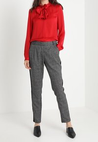 Blouse en satin rouge avec nœud au col associée à un pantalon gris à motifs fusel és avec des bandes noires sur les côtés, et des chaussures slip-on noires.