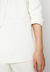 White blazer featuring a single chest pocket, with rolled-up sleeves and smooth fabric texture. Minimalist design with clean lines.