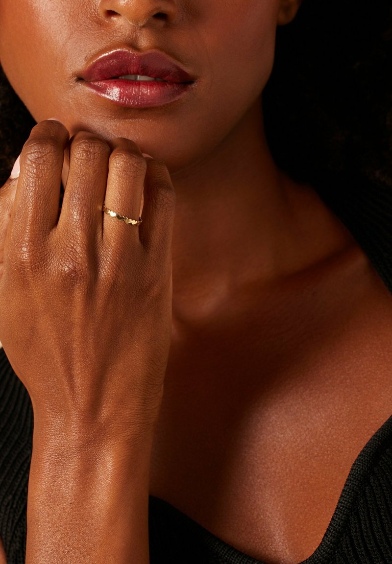 Gouden ring met een golvend patroon op een hand die naast het gezicht rust, met gladde huid en glanzende lippen. De achtergrond is donker.
