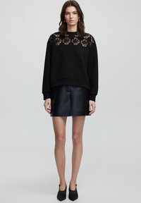 Maglione nero con dettagli in pizzo sulle spalle, abbinato a una mini gonna in pelle nera. L'outfit include scarpe nere a punta.